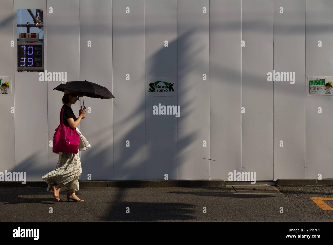 A woman using a parasol walks past a construction site wall with a noise pollution meter. Seijogakuenmae, Tokyo, Japan. Stock Photo