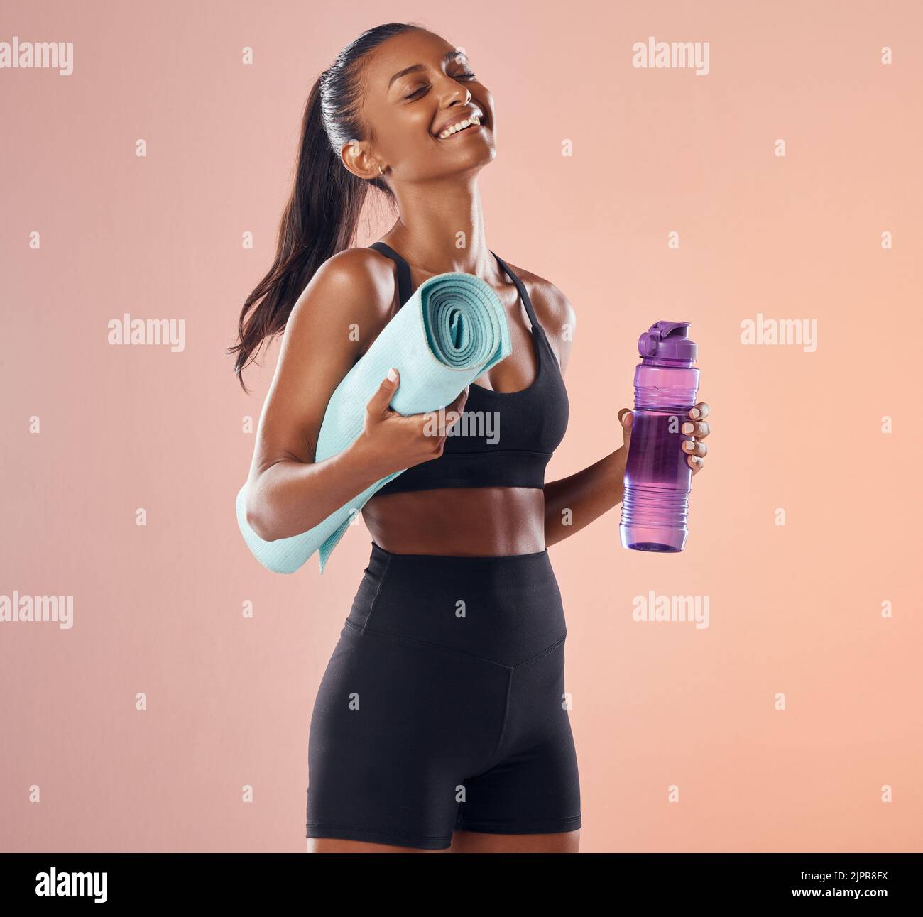Happy, fitness and healthy while holding exercise mat and water bottle against peach studio background. Fit woman with slim body after exercise Stock Photo