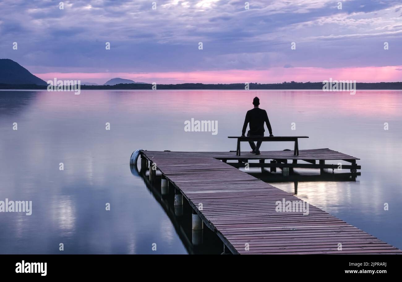 Man sitting on a wooden pier bench watching the sunrise at Lagoa da Conceicao in Florianopolis - Brazil Stock Photo