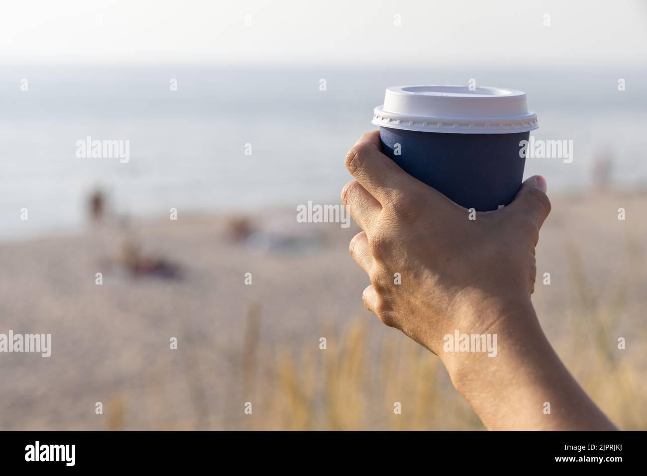 Young woman holds a disposable cup of coffee on on a sunny morning at the beach. Take away coffee at the sea. Stock Photo