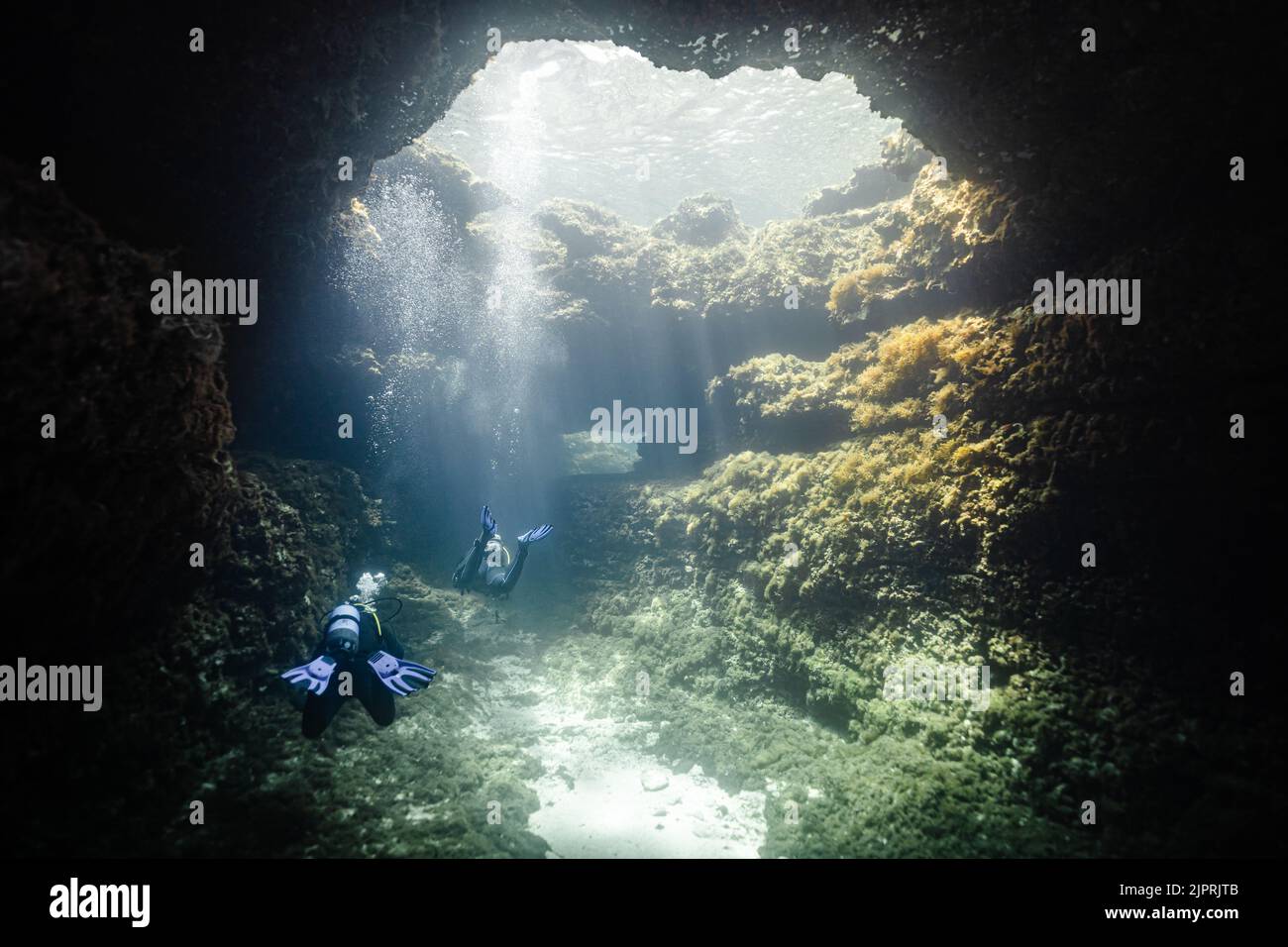 Two divers diving through cavern oh their way back from Alex Cave in Comino, Malta with light shining on them through a hole in a relaxed atmosphere. Stock Photo