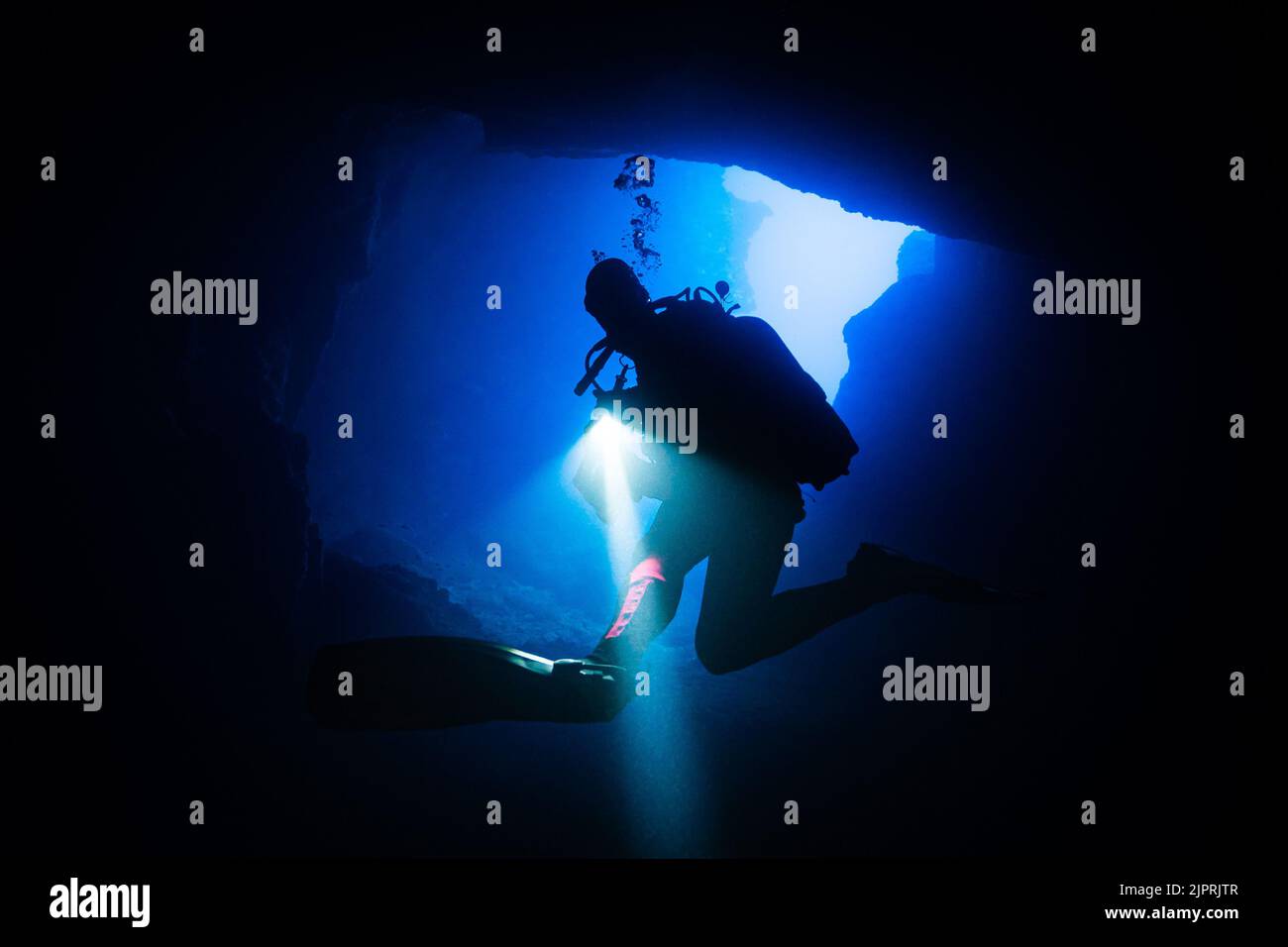 Diver turning around in front of a cave exit with shining flashlight in hand, while light shines in from outside in Blue Hole on Gozo, Malta. Stock Photo