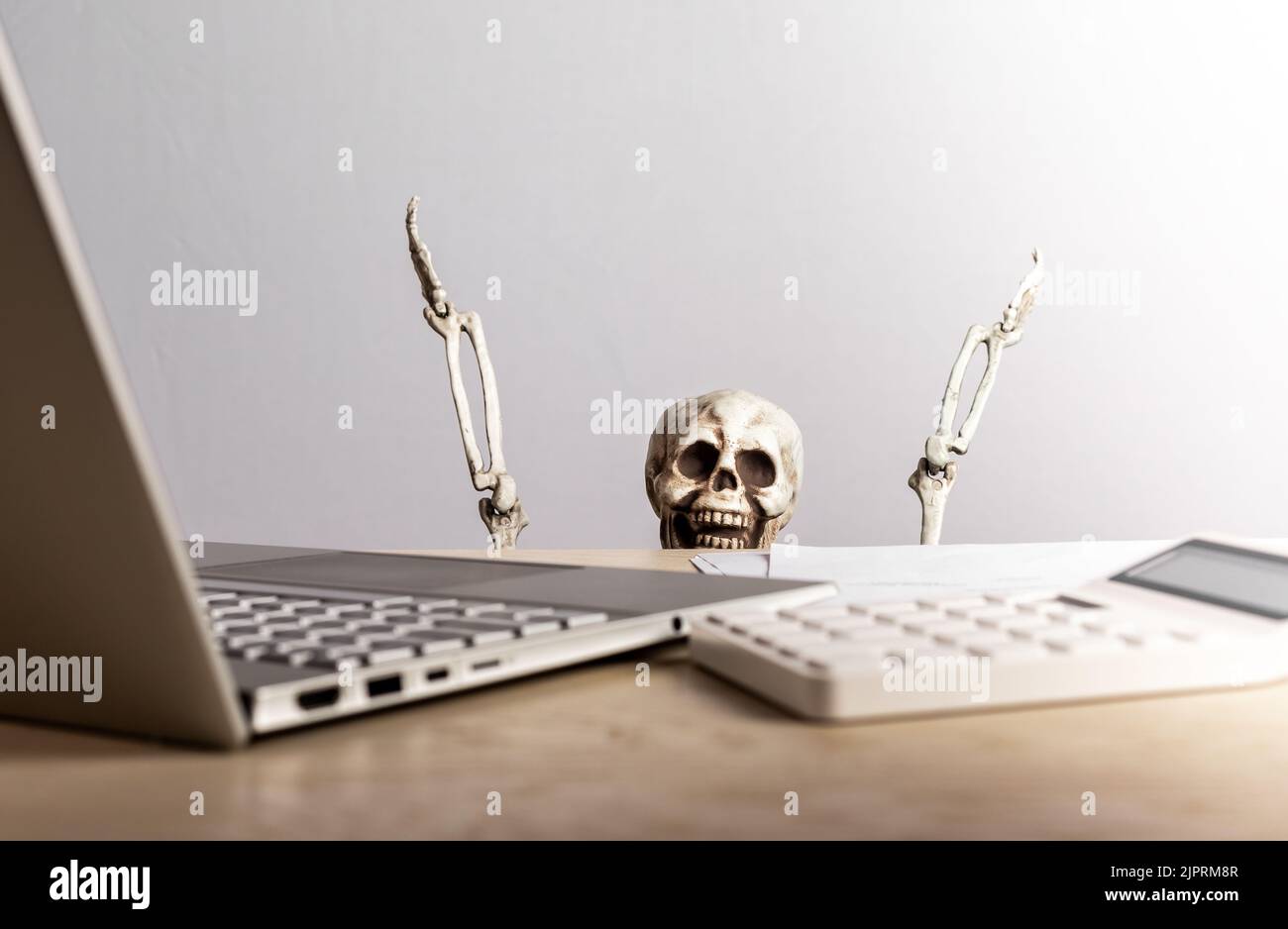 Job burnout. Skeleton peeking out from desk with laptop and calculator. Computer geek. Emotional and physical exhaustion, frustration, stress from wor Stock Photo
