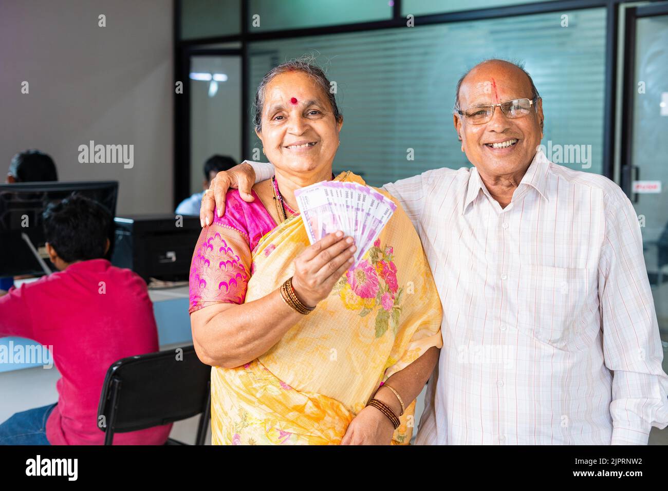 Happy smiling senior couple showing indian money or currency notes by looking camera at bank - concept of loan approval, investment, banking and Stock Photo
