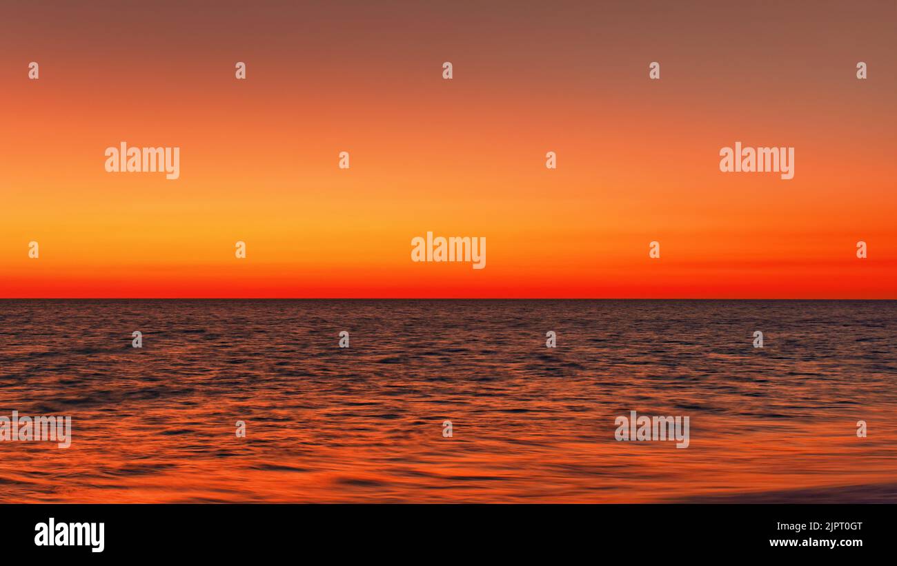 Vibrant orange red sky reflecting in calm ocean after sunset. Minimal landscape photo with space for text Stock Photo