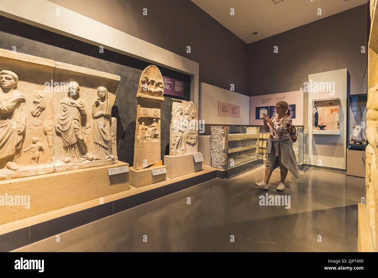 07.13.2022 Thessaloniki, Greece. Curious caucasian middle-aged female tourist walking past ancient Greek sculptures and statues and taking a photo with her smartphone. High quality 3d illustration Stock Photo