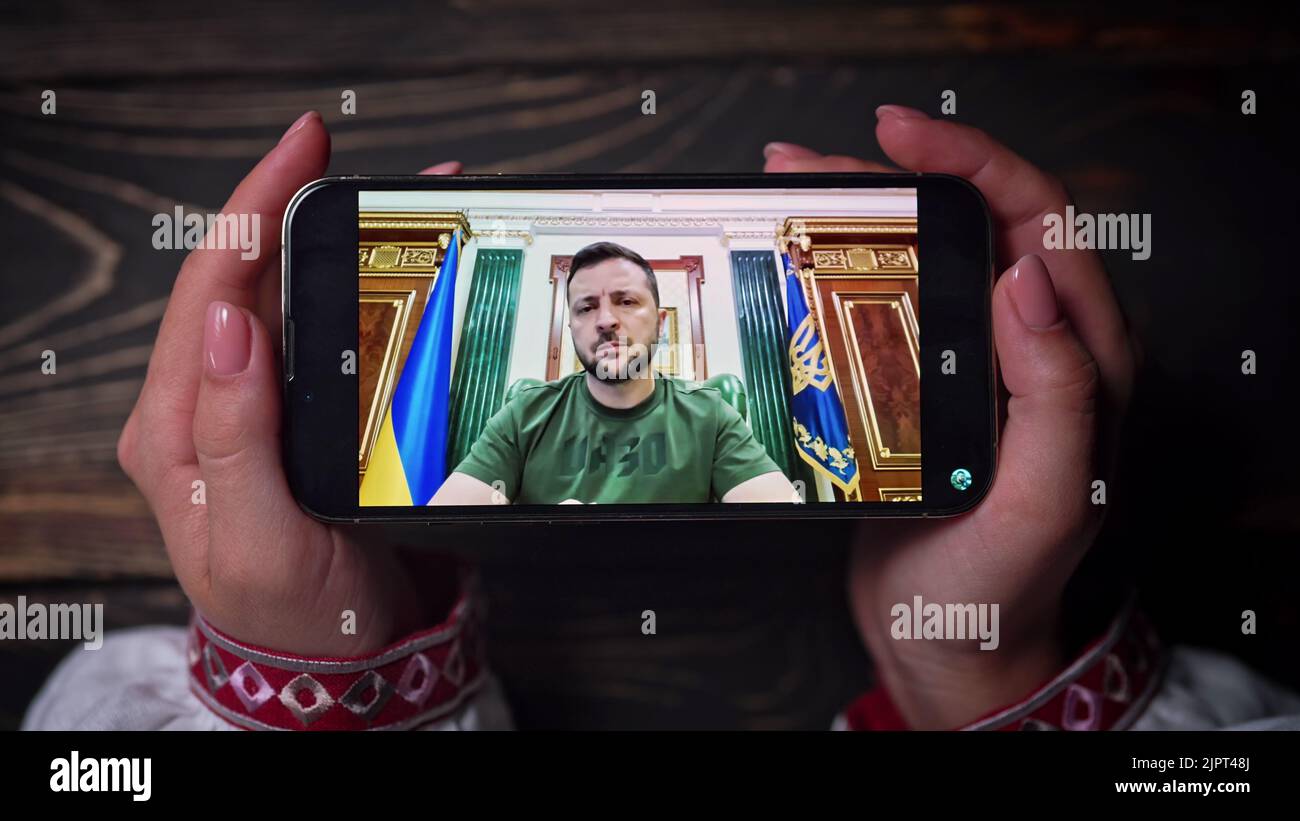August 2022 - Kyiv, Ukraine. Watching online speech of President Volodymyr Zelenskiy on smartphone on wooden table background. News from front Stock Photo