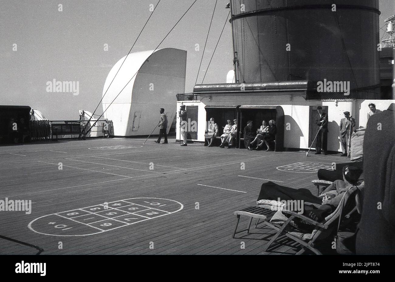 1950s, historical, passengers on a steamship, some holding long poles, playing a deck game, shuffle-board, other travellers sitting in little covered shelters watching the play. At the side of the deck, passengers lying in wooden lounger chairs with cunard embrodied chair blankets. Stock Photo