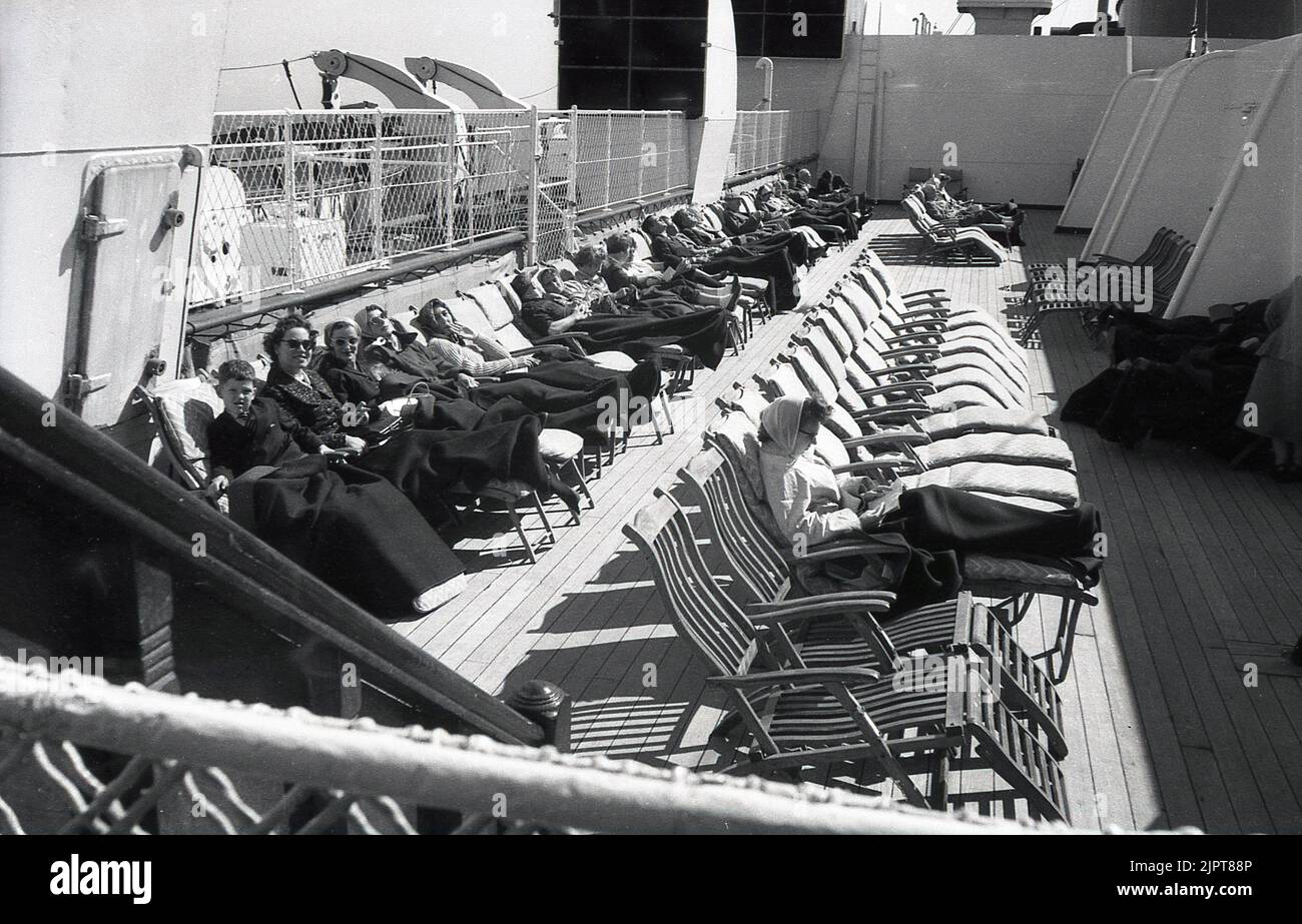 1950s, historical, outside on a cruise ship of the era, on a Cunard White Star liner, on a section of the steamship, passengers, most with coats on and woollen chair blankets over their legs, relaxing, sitting in the wooden deck loungers, enjoying the sunshine. Stock Photo