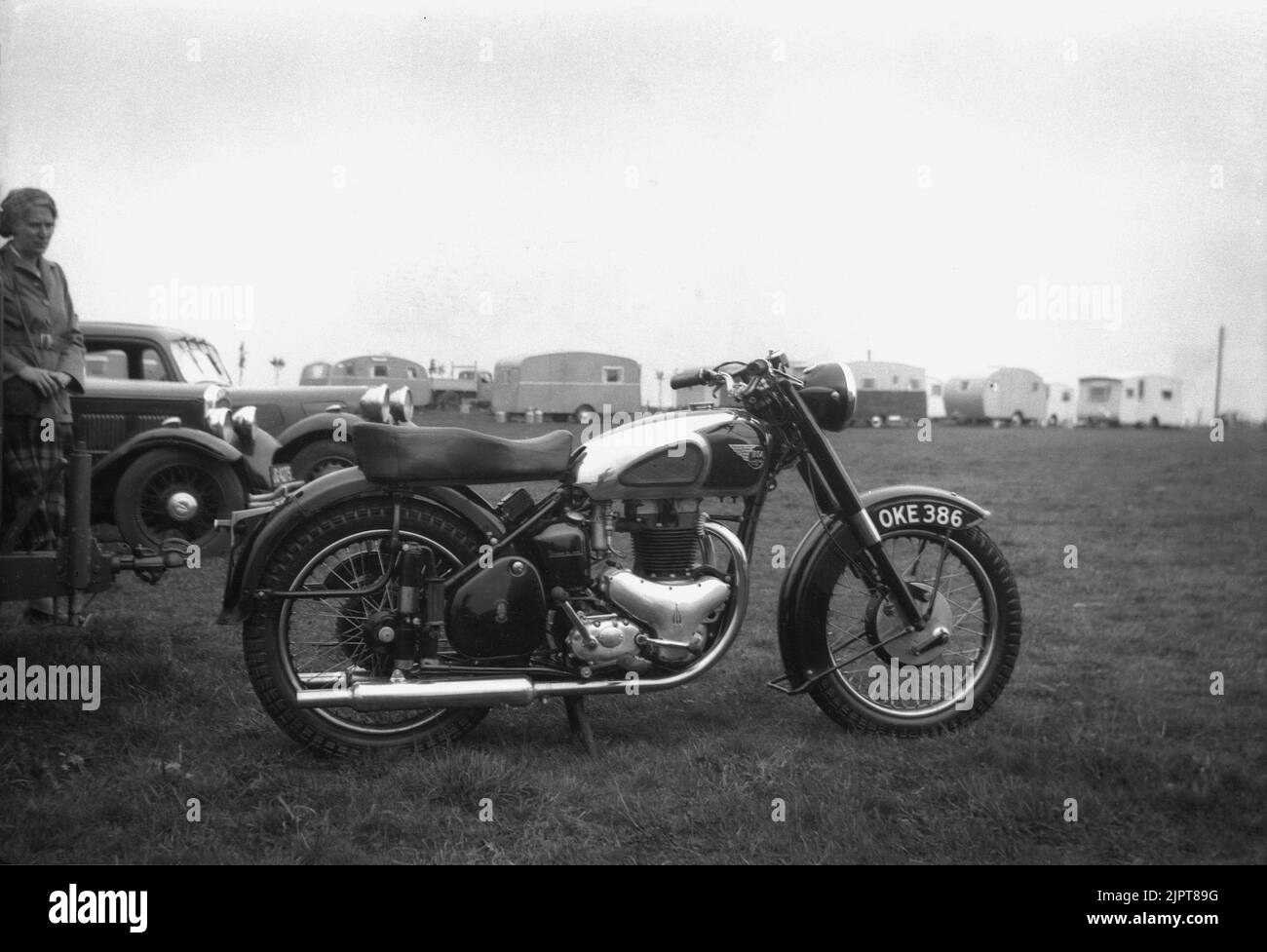 1950s, historical, outside in a field at a caravan site, a lady, standing by some motorcars looking at a British made BSA motorcycle of the era parked on the grass, England, UK. Founded in 1861 as the Birmingham Small Arms Company Ltd, for the production of firearms, a motorcycle division of BSA was established in 1903, and the first motorcycle released in 1910. In the 1950s, BSA Motorcycles was the largest motorcycle maker in the world. Stock Photo