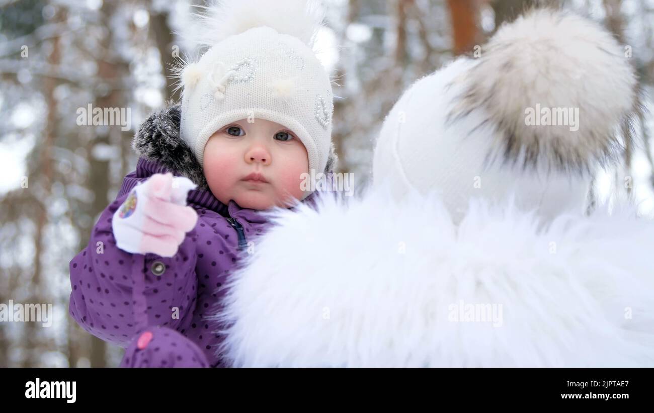 on the background of the winter forest, a portrait of a small one-year-old girl in a funny hat with a bumbon. the girl reaches into the camera with her hand. High quality photo Stock Photo