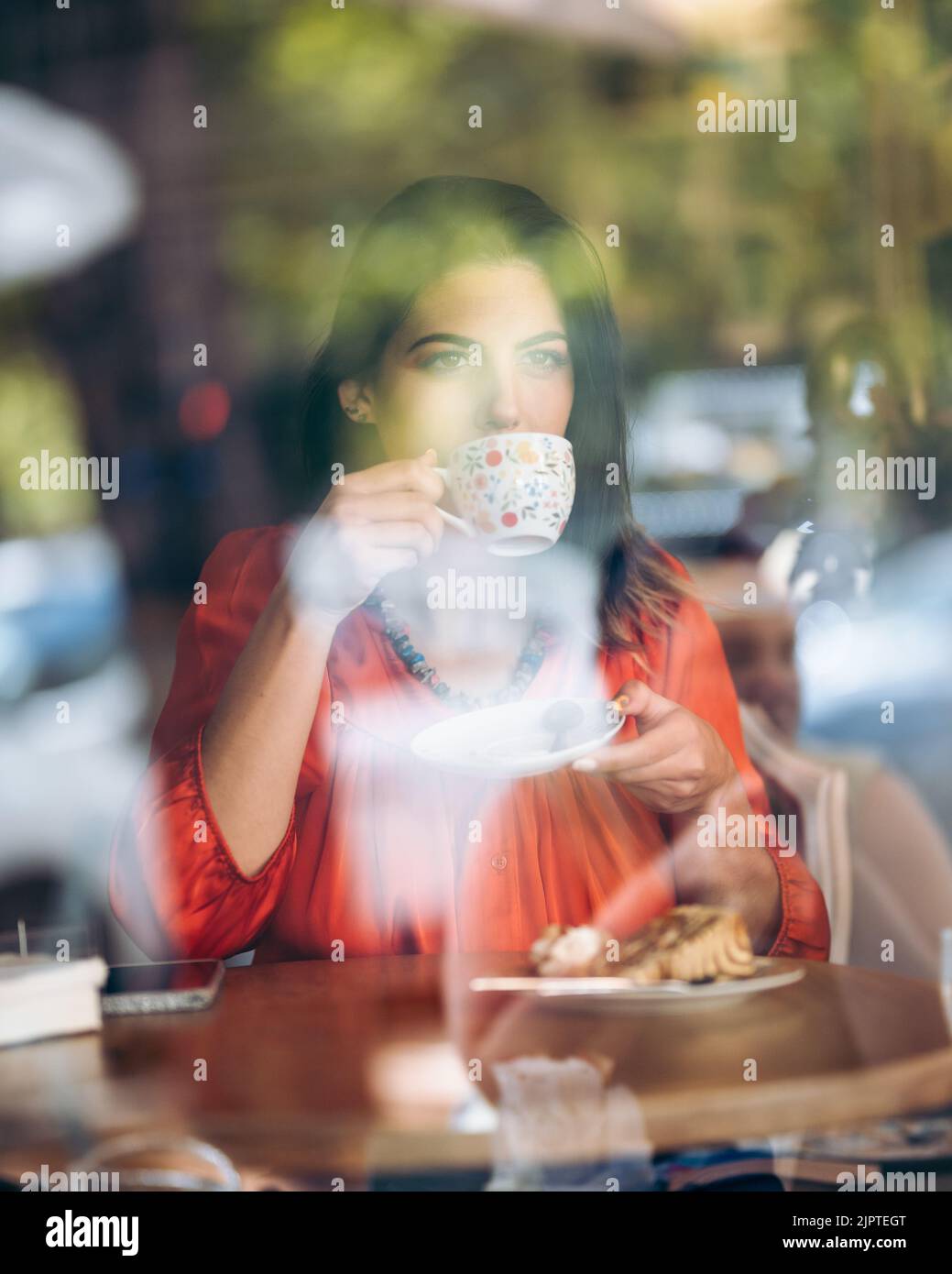 Pensive young woman drinks coffee while looking out the window seen ...