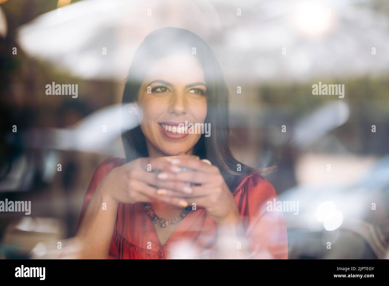Smiling young woman drinks coffee while looking out the window seen ...