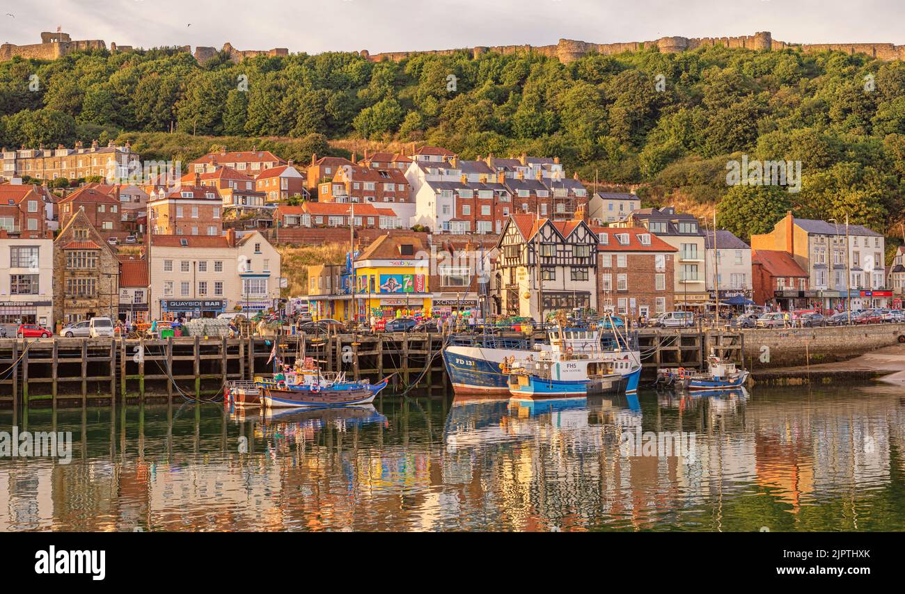Fishing boats are moored to a quay with their reflections in the water. Cafes and shops line the seafront.  The hill behind is topped by the wall of a Stock Photo