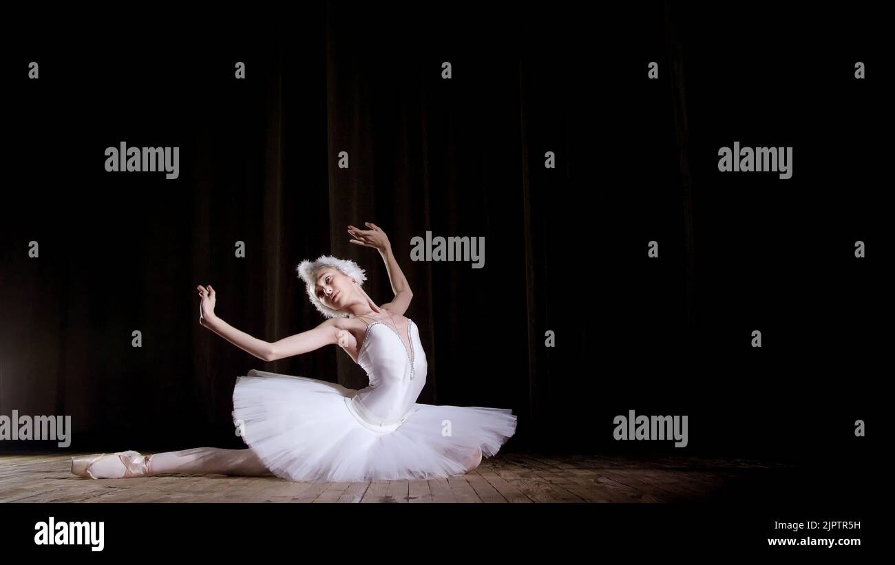 in rays of spotlight, on the stage of the old theater hall. Young ballerina in suit of white swan and pointe shoes, dances elegantly certain ballet motion, Swan Lake. High quality photo Stock Photo