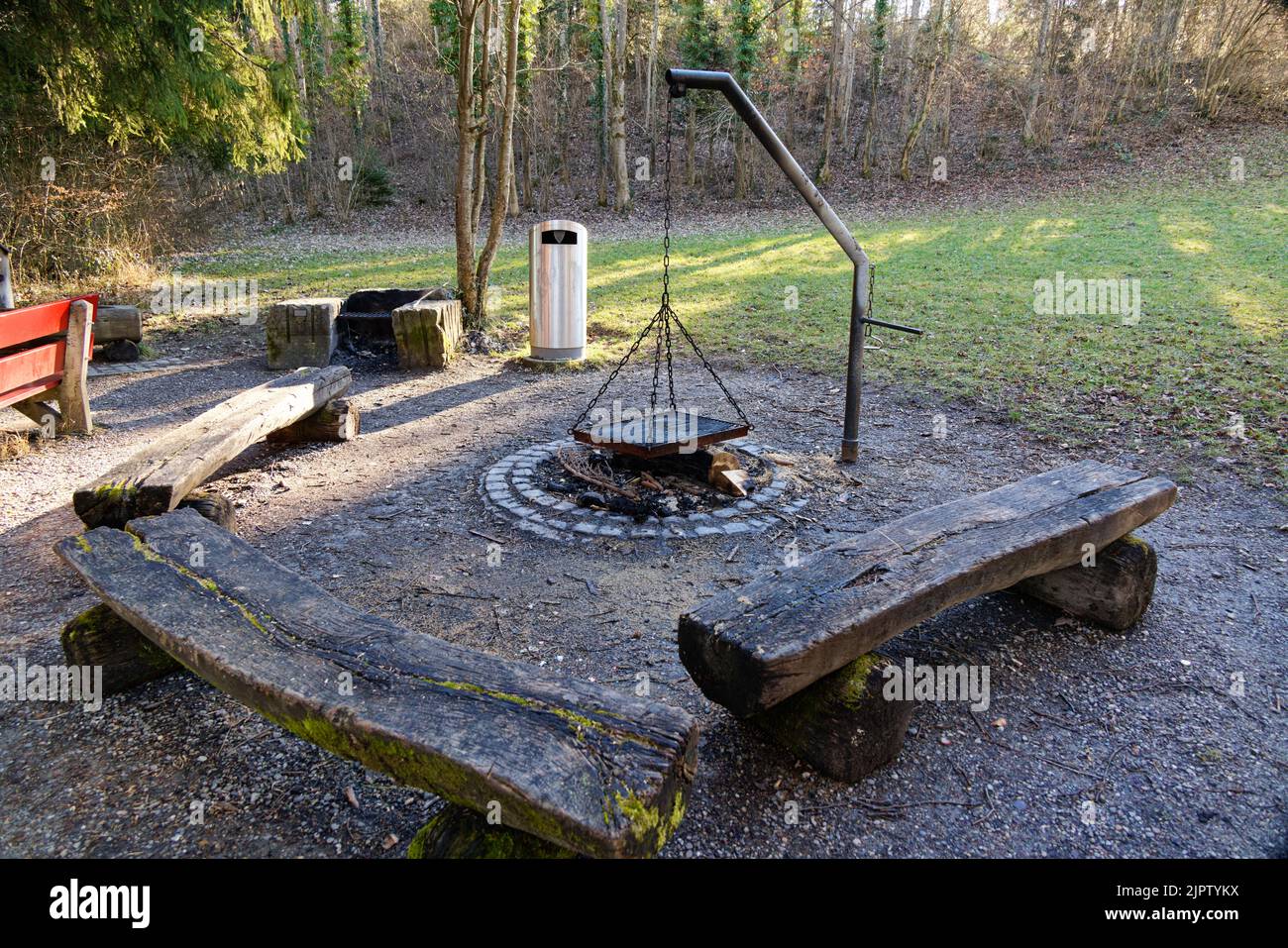 large barbecue area with metal grill and many wooden benches, in the shade, fire is extinguished, trash can in the background, green meadow, in front Stock Photo