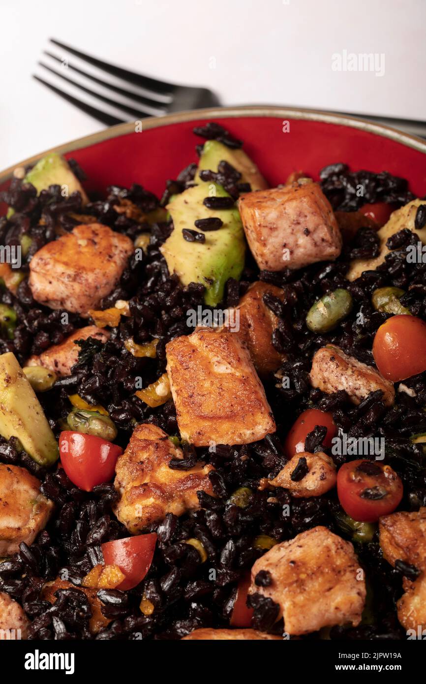 A red bowl of black rice with baked salmon, avocado, tomatoes, chili
