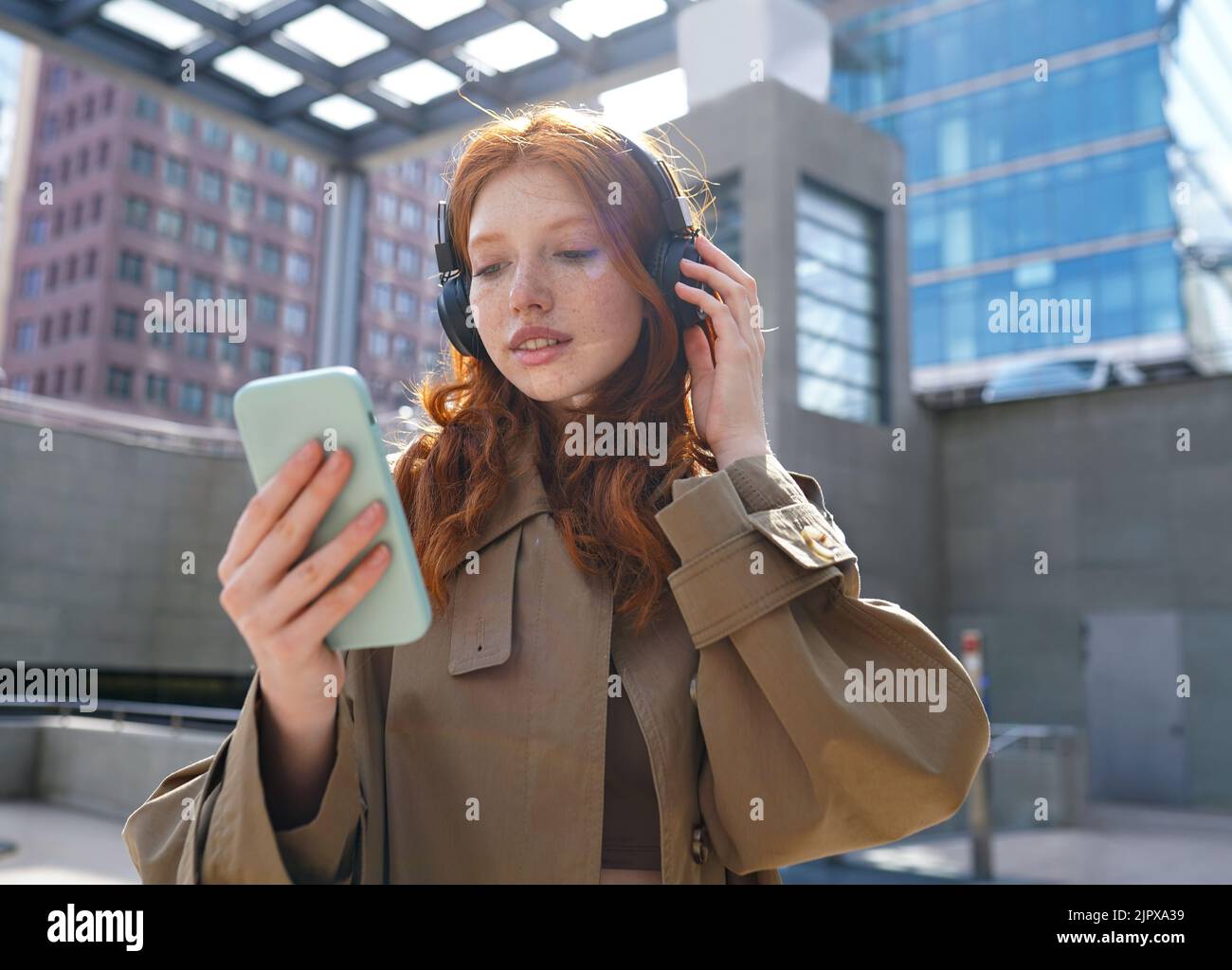 Teen redhead girl wearing headphones using smartphone in big city. Stock Photo