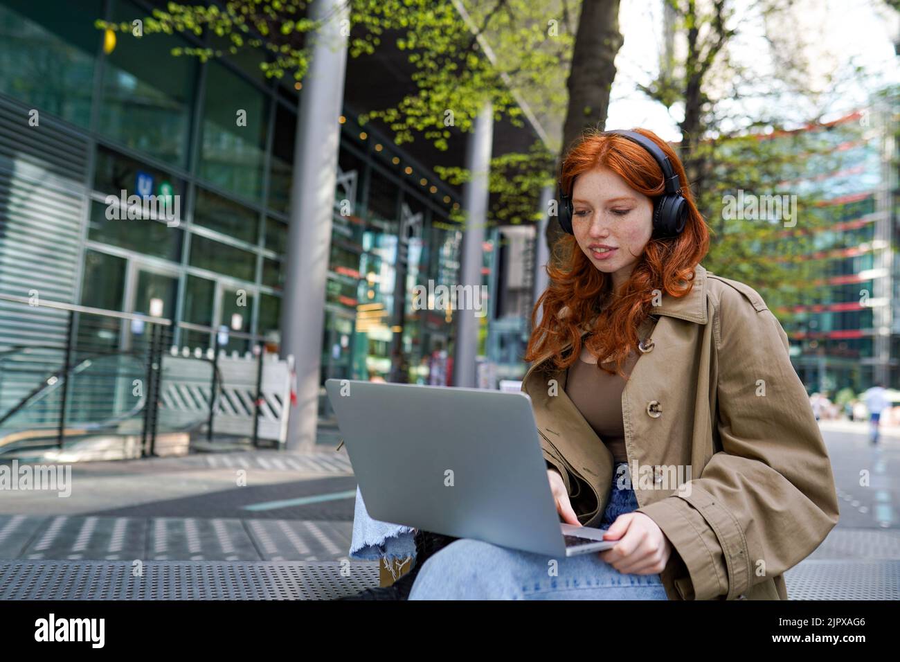 Teen redhead hipster girl wearing headphones using laptop on city urban street. Stock Photo