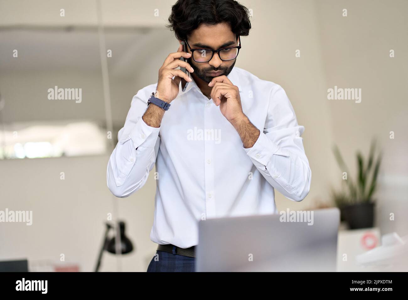 Busy indian business man talking on phone looking at laptop in office. Stock Photo
