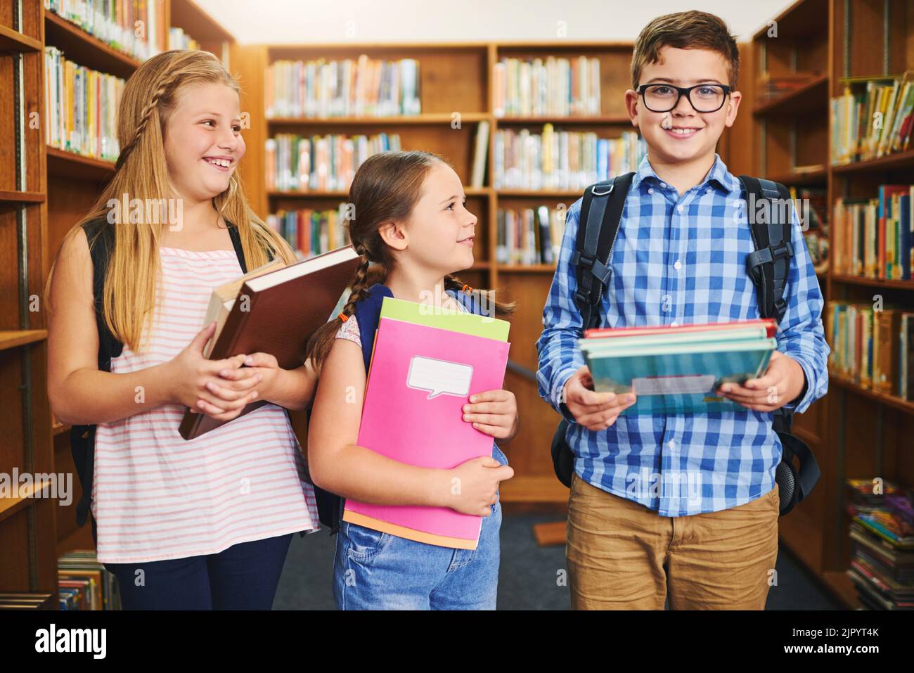 The girls are crazy about him. a group of young school kids holding books while standing inside of a library together during the day. Stock Photo