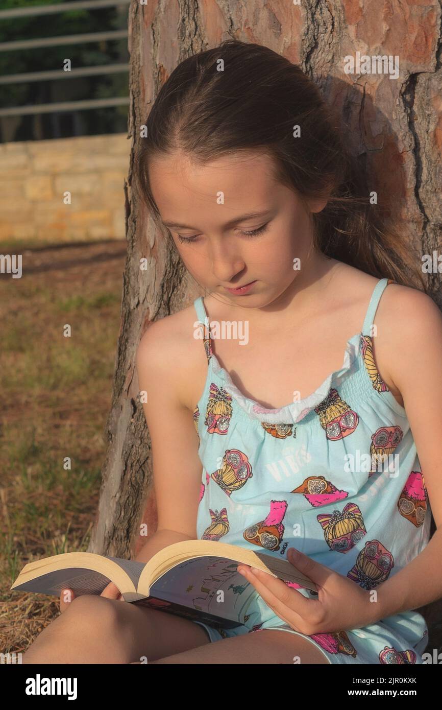 The little girl reading a book while sitting under a tree in the park ...