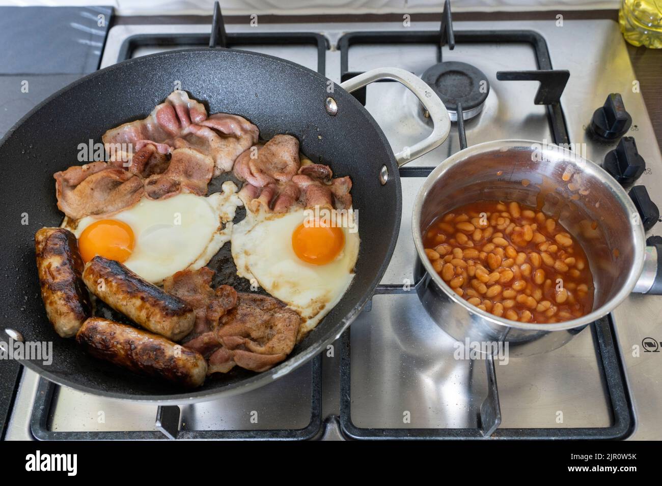 Traditional English breakfast ingredients cooking and frying on a gas hob. Bacon, eggs, sausages and baked beans.UK. Concept - fatty meal Stock Photo