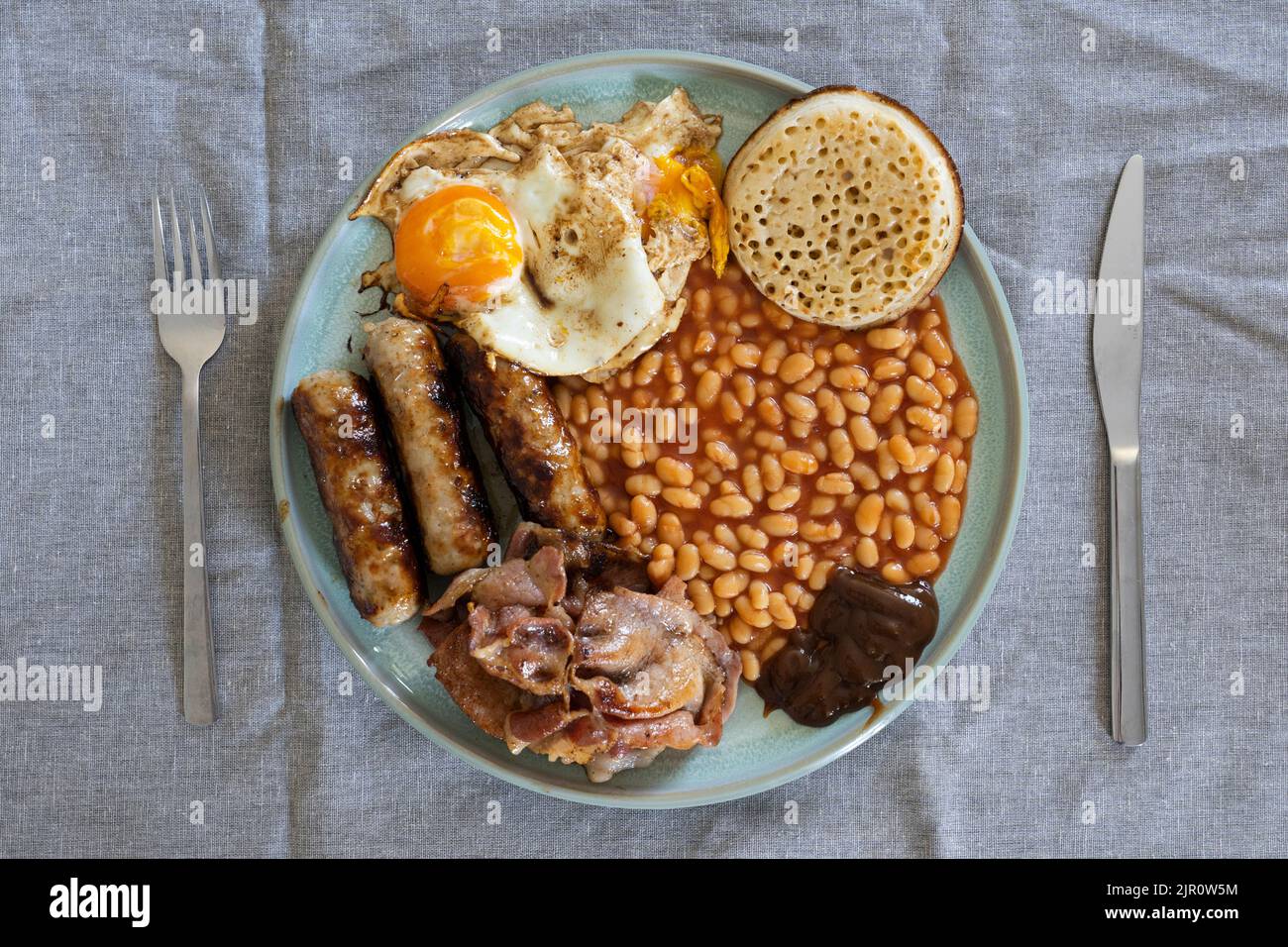 Traditional English breakfast on a table with cutlery - eggs, bacon, sausages, baked beans and a crumpet. UK. Concept - unhealthy eating Stock Photo
