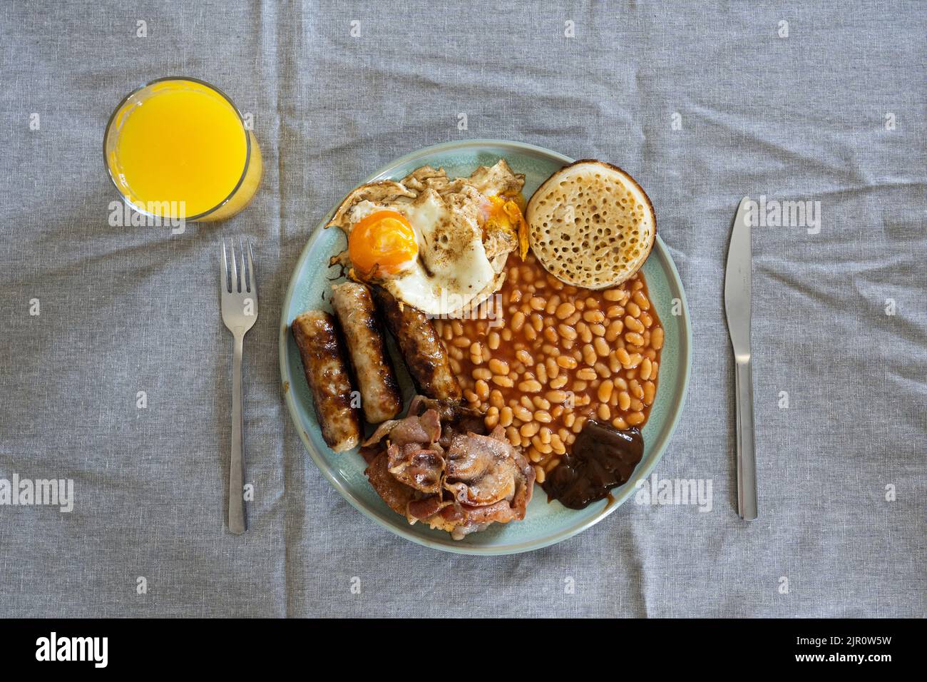 Traditional English breakfast on a table with orange juice, cutlery, eggs, bacon, sausages, baked beans and a crumpet. UK. Concept - unhealthy eating Stock Photo