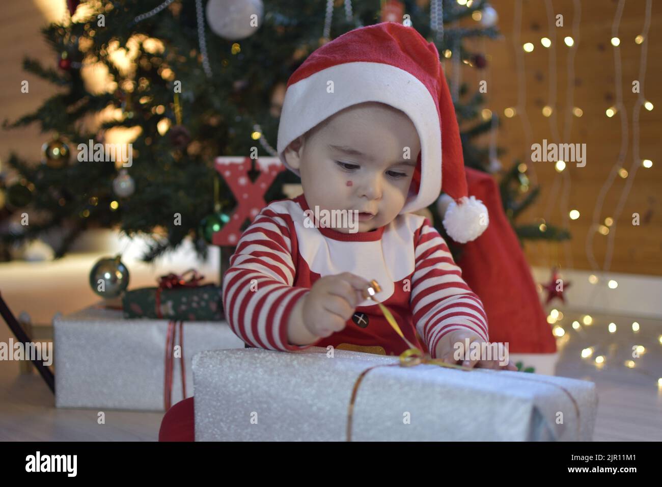 Twelve months old baby opening a Christmas gift. baby dressed as santa claus opening gift. cute baby next to the christmas tree.excited to open gift Stock Photo