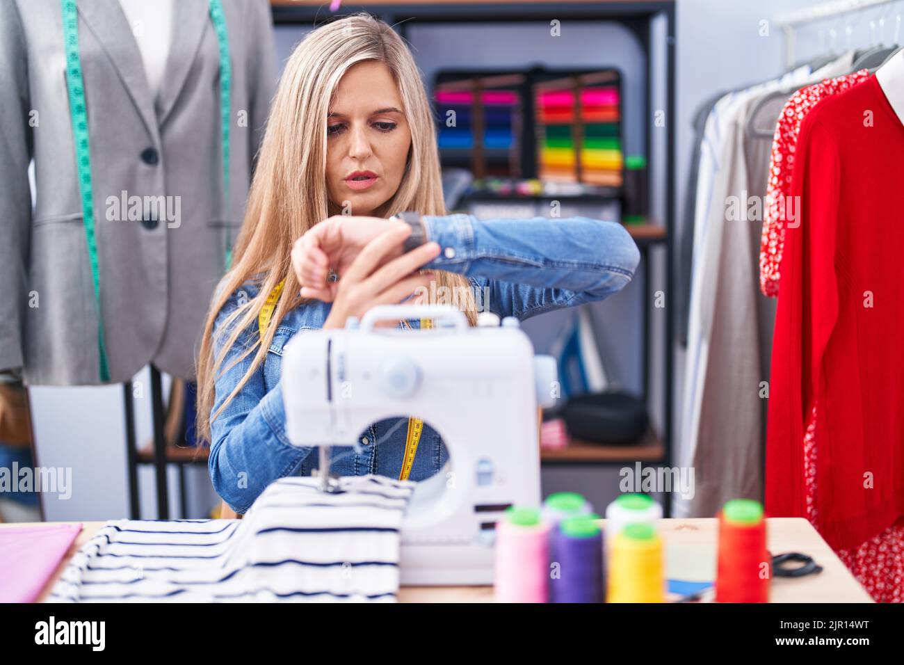 Blonde woman dressmaker designer using sew machine checking the time on wrist watch, relaxed and