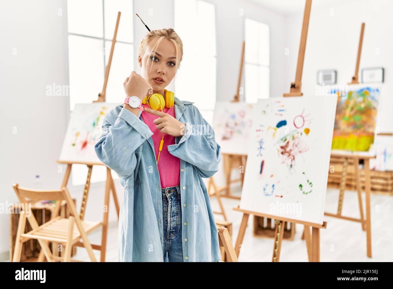 Young caucasian girl at art studio in hurry pointing to watch time ...