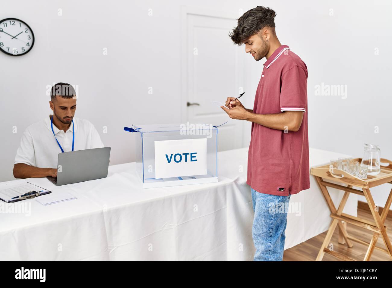 Young hispanic voter man smiling happy writing on vote at electoral center. Stock Photo