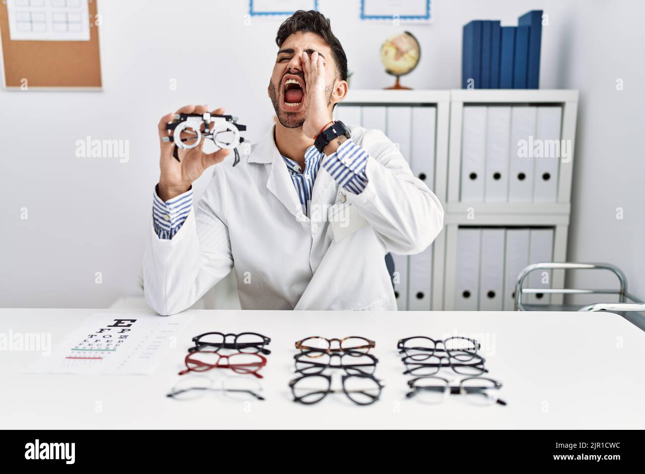 Young optician man holding optometry glasses shouting and screaming
