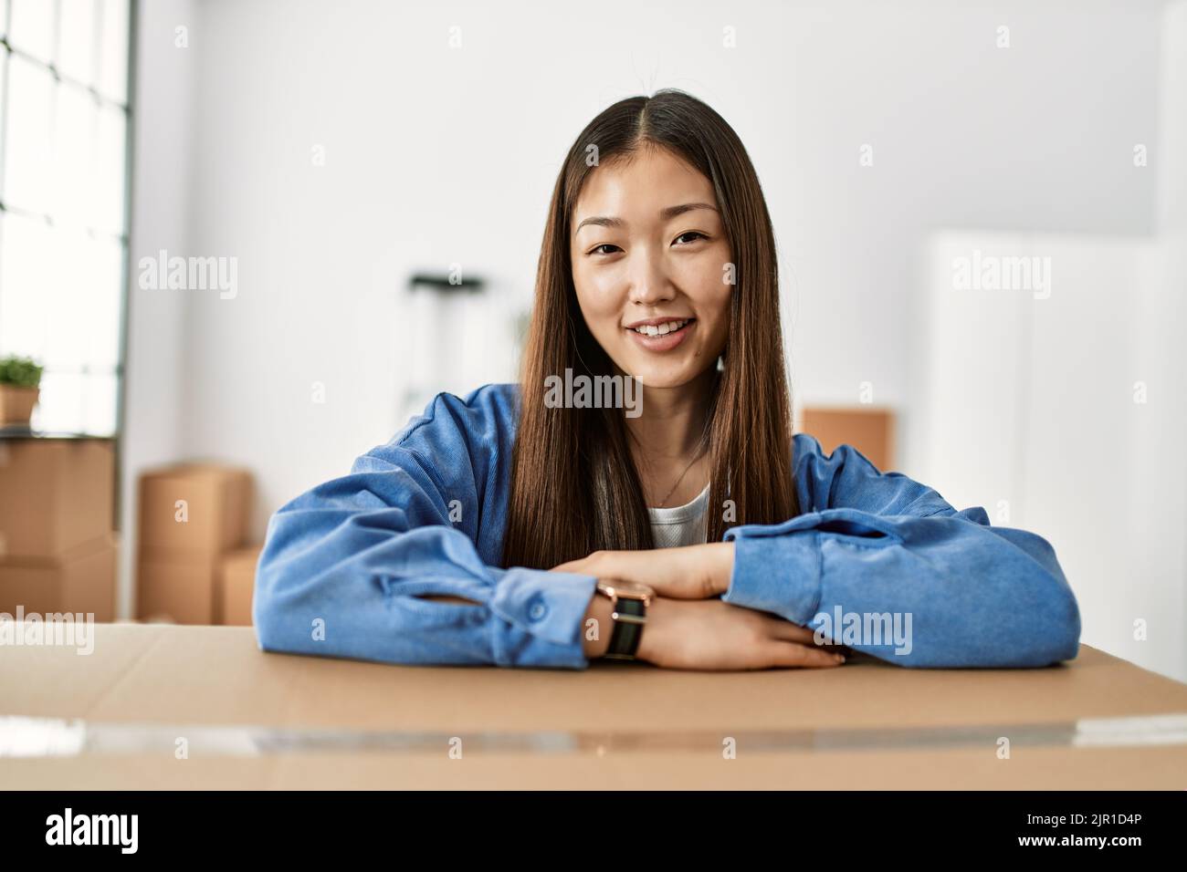 Young chinese girl smiling happy leaning on cardboard box at new home ...