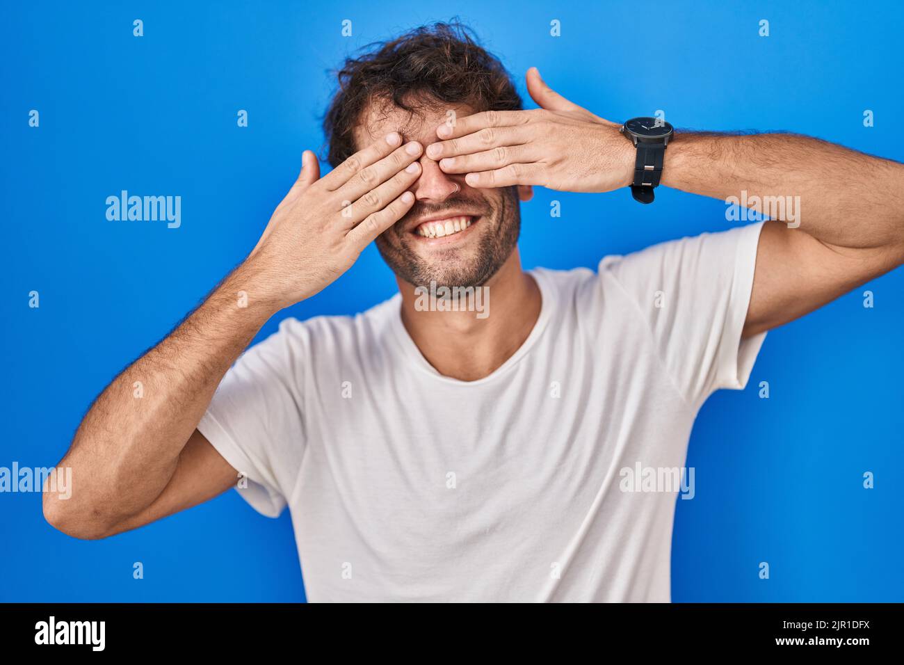 Hispanic young man standing over blue background covering eyes with ...
