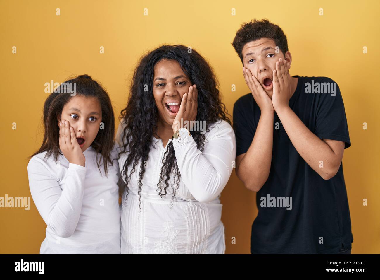 Family of mother, daughter and son standing over yellow background afraid and shocked, surprise and amazed expression with hands on face Stock Photo