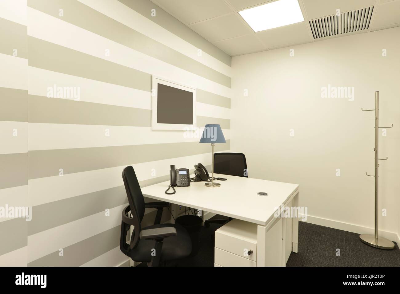 Office cubicle with small white double table with telephones, black swivel chairs, metal coat rack, scratched wall and dark gray carpet floor Stock Photo