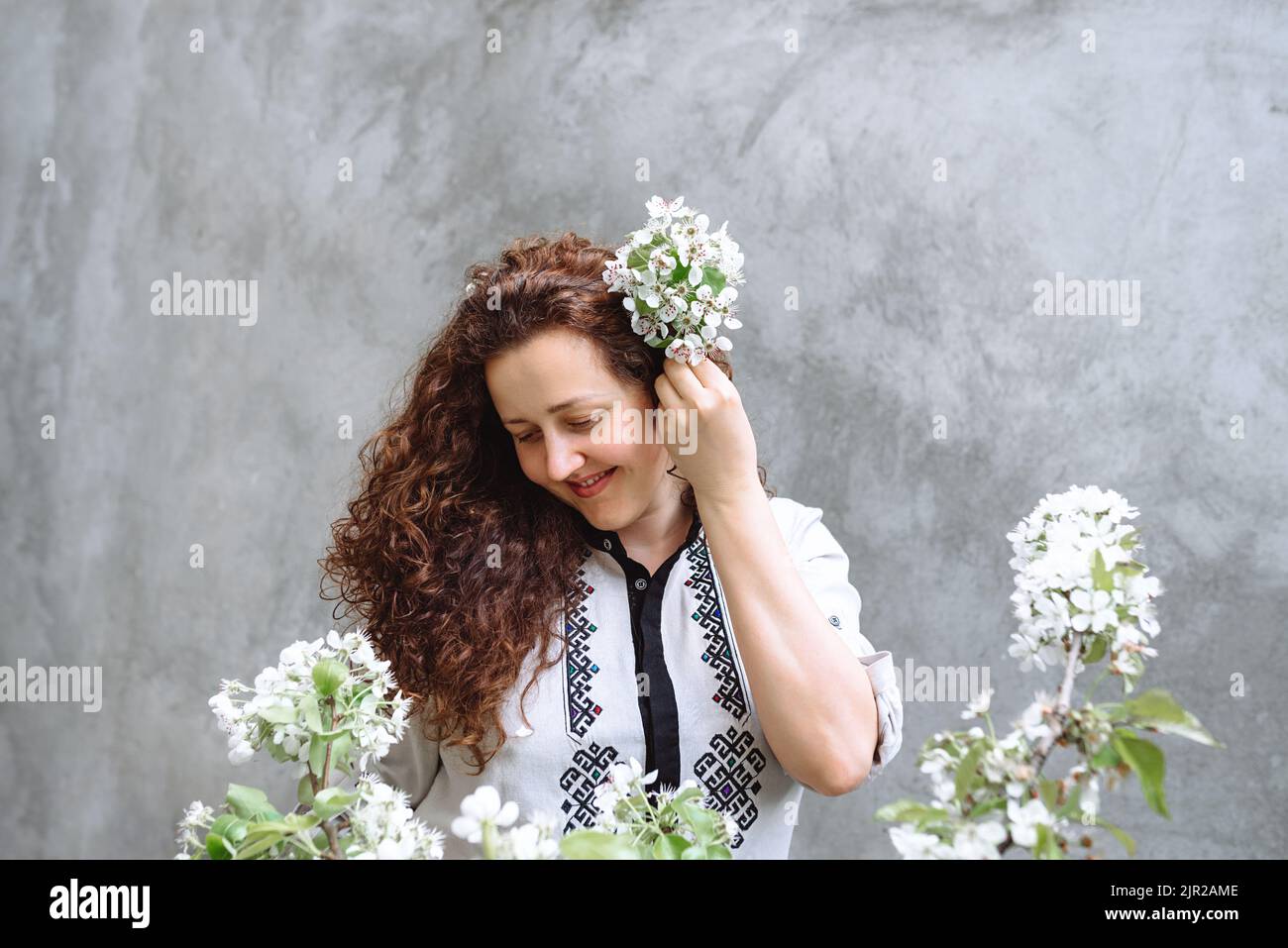 Girl with curly hair in a shirt with a downcast look. Woman holds blossoming branch of apple tree in hand against background of concrete wall. Spring flowering time. Constitution Day of Ukraine Stock Photo