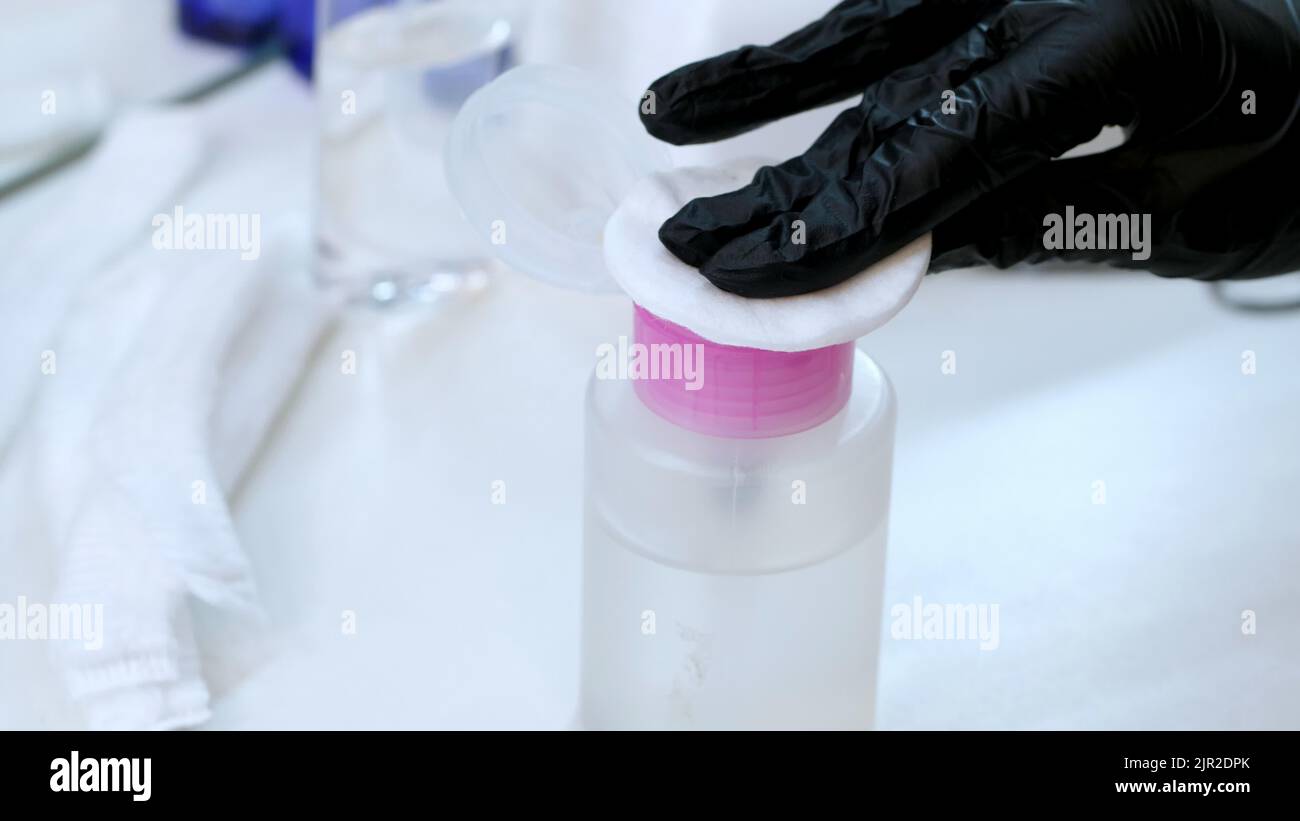 close-up, the hands of the beautician in black rubber gloves hold the cotton pad and moisten it in a special transparent liquid from the bottle with a dispenser. High quality photo Stock Photo