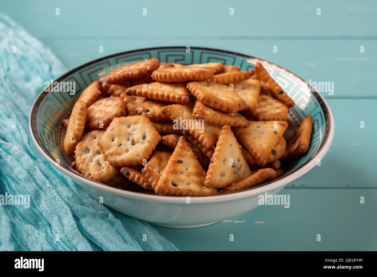 Teal bowl full of saltine crackers over turquoise wood background. Salty crackers with black and white sesame seeds in a blue deep plate. Ready to eat Stock Photo