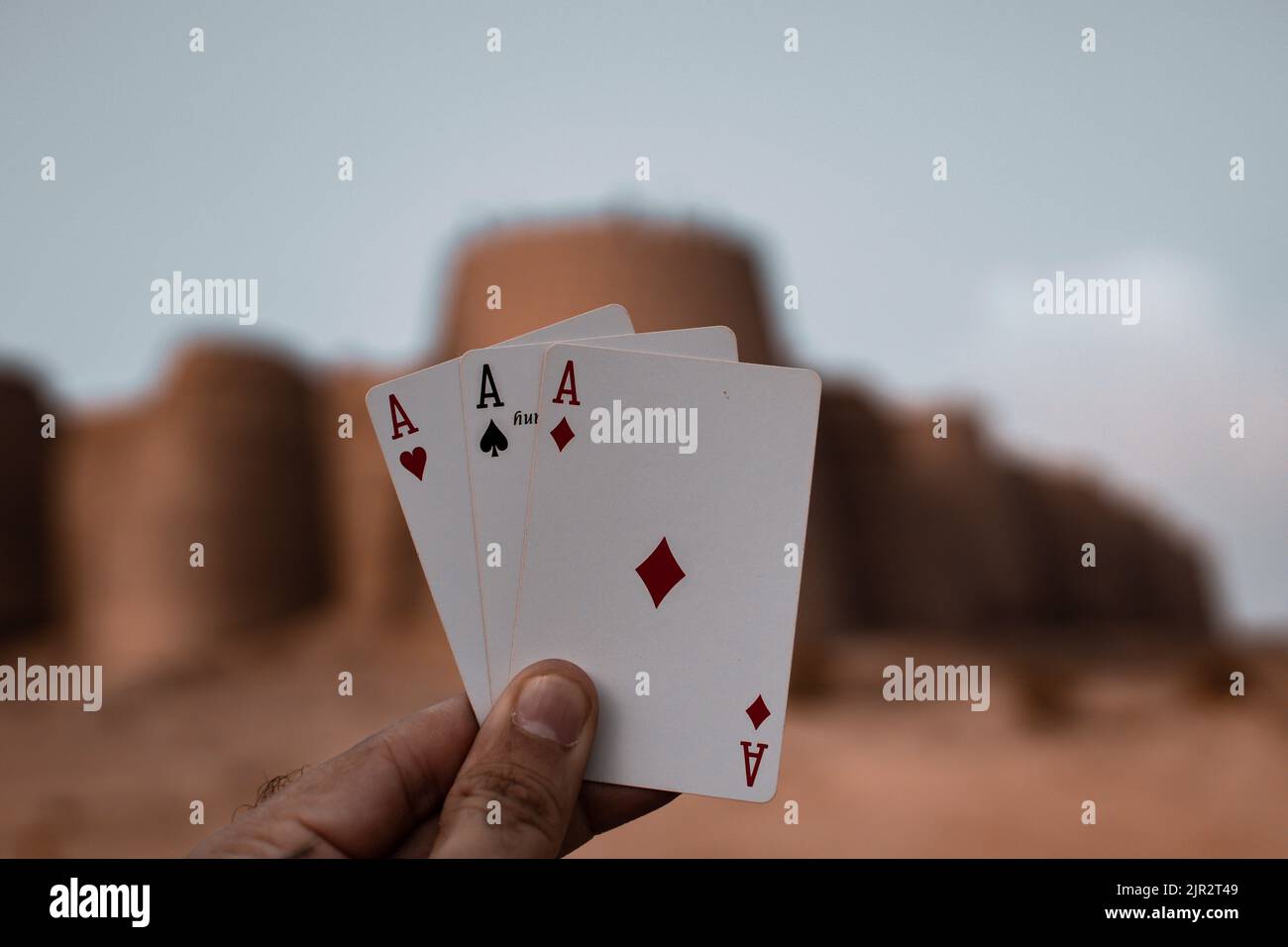 A close-up shot of a hand holding three aces in the background of the Derawar Fort in Pakistan Stock Photo