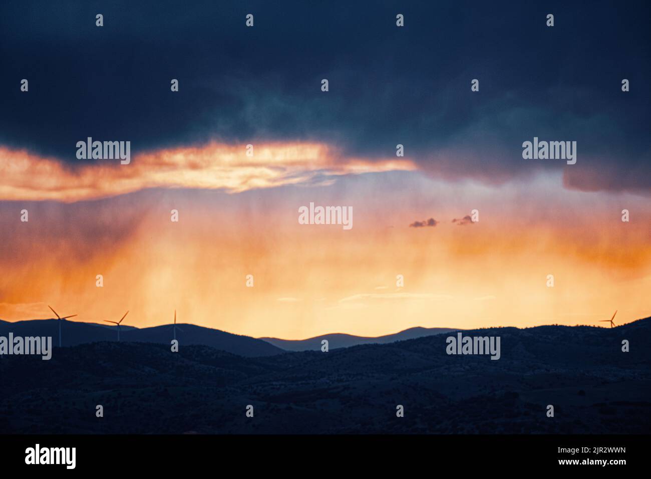 Dramatic sunset storm clouds with rainfall over rolling hills and distant wind turbines Stock Photo