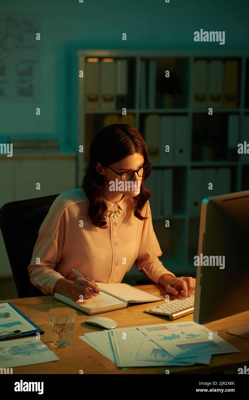 Young woman reading data on computer screen and taking notes Stock Photo