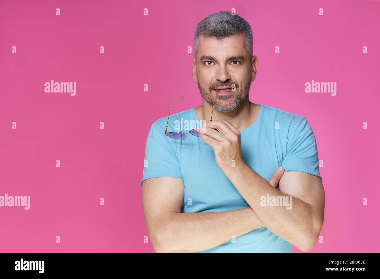 Handsome adult man with grey hair in casual isolated on pink background. Good-looking middle aged man standing with arms folded holding sunglasses and smile wearing blue t-shirt.  Stock Photo