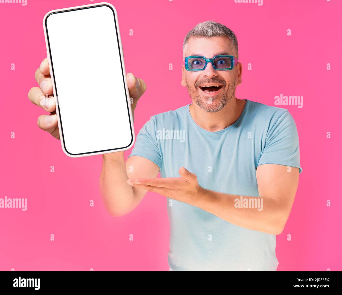 Happy excited middle aged handsome man 40s with big smartphone in hand wearing blue shirt and sunglasses isolated on pink background. Mobile app advertisement.  Stock Photo