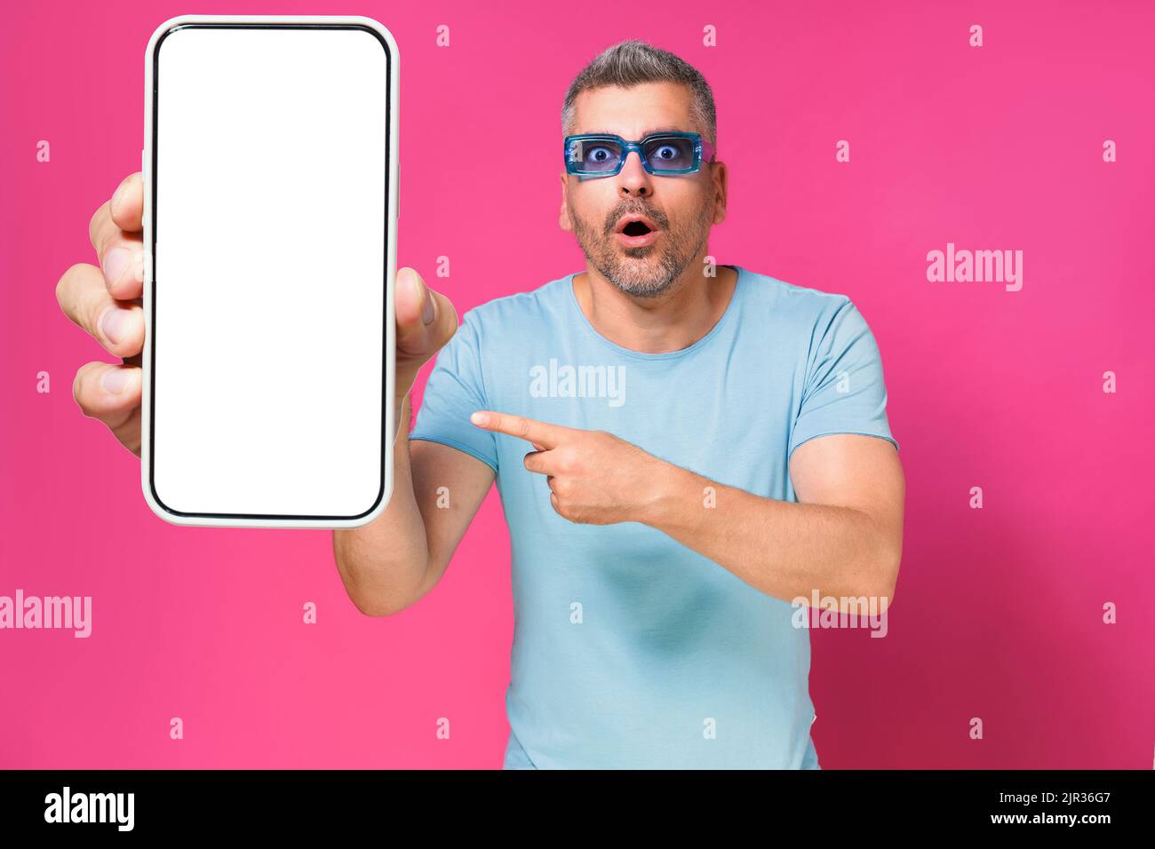 Shocked handsome man 40s pointing at big smartphone in hand wearing blue shirt and sunglasses isolated on pink background. Man with phone studio shot. Mobile app advertisement. Copy space. Stock Photo