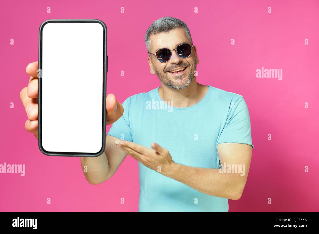 Mobile app advertisement on smartphone screen in hand of handsome man, guy 30s 40s in casual blue shirt and sunglasses isolated on pink background. Man with phone studio shot.  Stock Photo