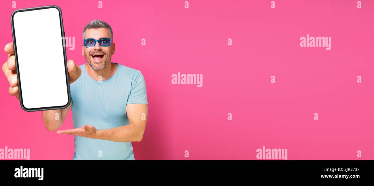 Big, huge smartphone in hand of handsome man, guy 30s 40s in casual blue shirt and sunglasses isolated on pink background. Man with phone studio shot. Mobile app advertisement. Copy space. Stock Photo