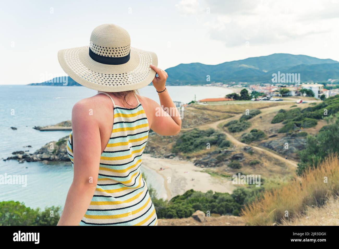Medium closeup shot of a young caucasian female tourist in sleeveless striped dress standing backwards to the camera, holding her beige summer hat so that the strong wind will not blow it away, and enjoying seashore view. Summer holidays and traveling concept. High quality photo Stock Photo